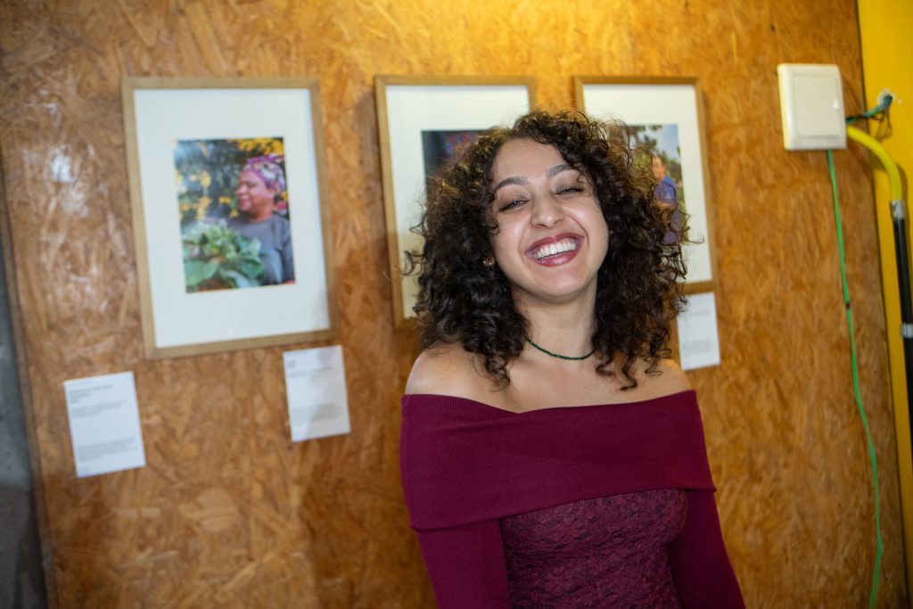 Mulher de cabelos cacheados escuros sorri amplamente para a câmera em um espaço expositivo. Ela veste uma blusa vinho de ombros à mostra e usa um colar fino verde. Ao fundo, há uma parede de madeira com fotografias emolduradas penduradas, compondo uma exposição. A iluminação é quente e destaca a expressão alegre e descontraída da pessoa retratada.